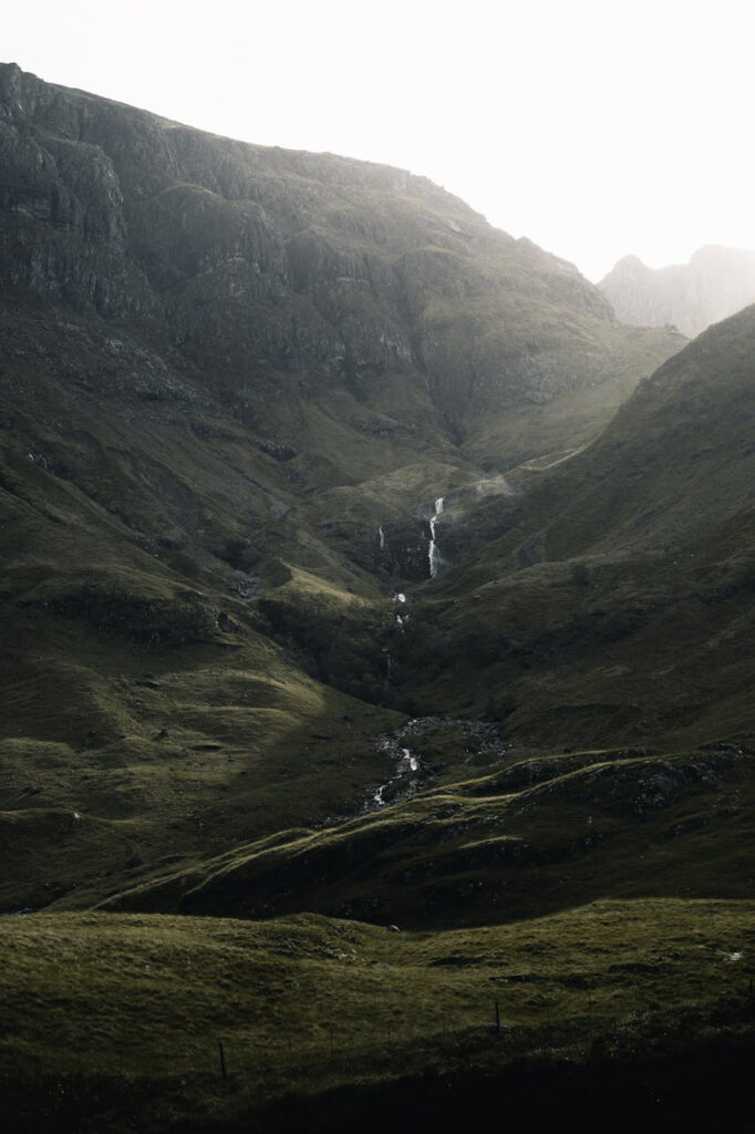 A serene landscape in Glencoe, featuring steep, rugged green hillsides with a cascading waterfall cutting through the valley, illuminated softly by diffused sunlight.