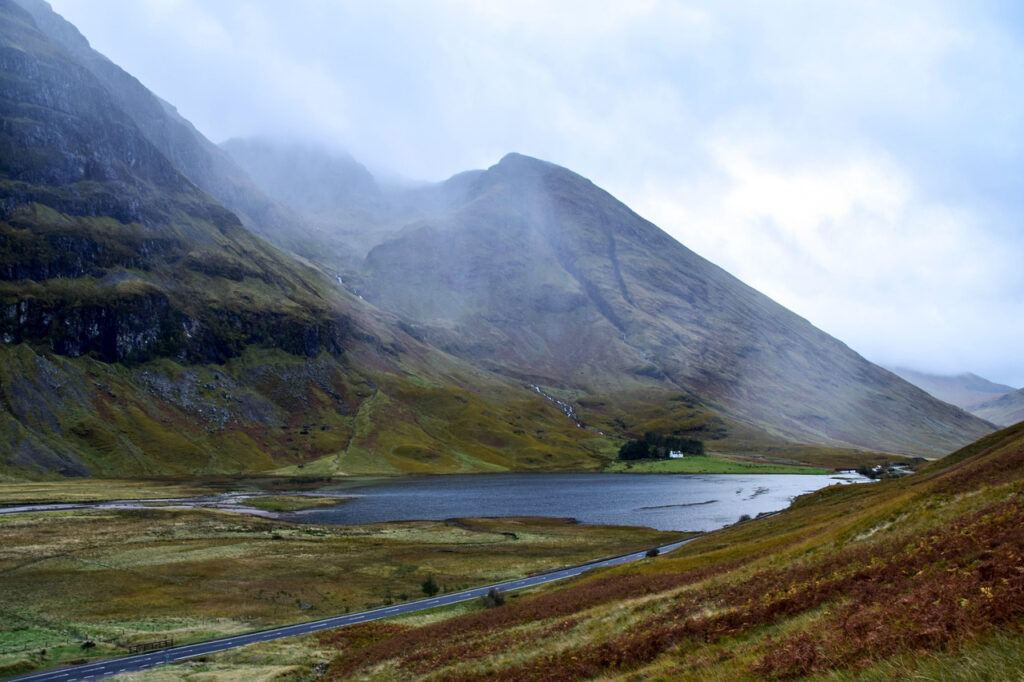 A serene landscape in Glencoe, featuring a tranquil loch surrounded by steep, mist-covered hills, with a winding road cutting through the foreground and a solitary white house nestled in the distance.