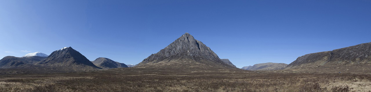 A panoramic view of Buachaille Etive Mor mountain in Glencoe, Scotland, surrounded by an expansive moorland under a clear, vibrant blue sky.