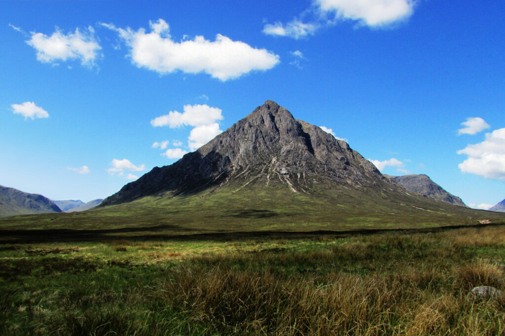 The iconic Buachaille Etive Mor mountain in Glencoe, Scotland, under a bright blue sky with scattered white clouds, surrounded by a vast, green moorland.