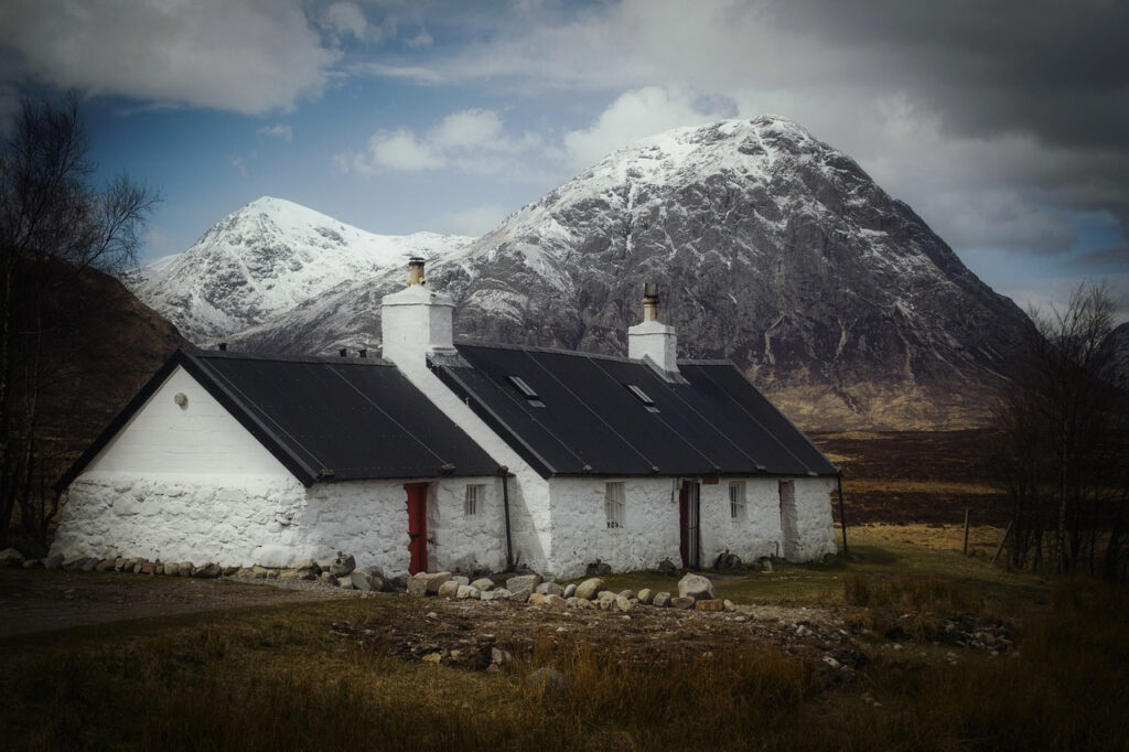 A traditional whitewashed cottage with a black roof, nestled in a remote moorland setting, with the snow-capped Buachaille Etive Mor mountain towering in the background under a cloudy sky in Glencoe, Scotland.