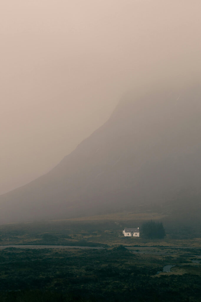 A solitary white house sits amidst a vast moorland under a dense layer of fog, with the faint silhouette of Buachaille Etive Mor mountain looming in the background, located in Glencoe, Scotland.