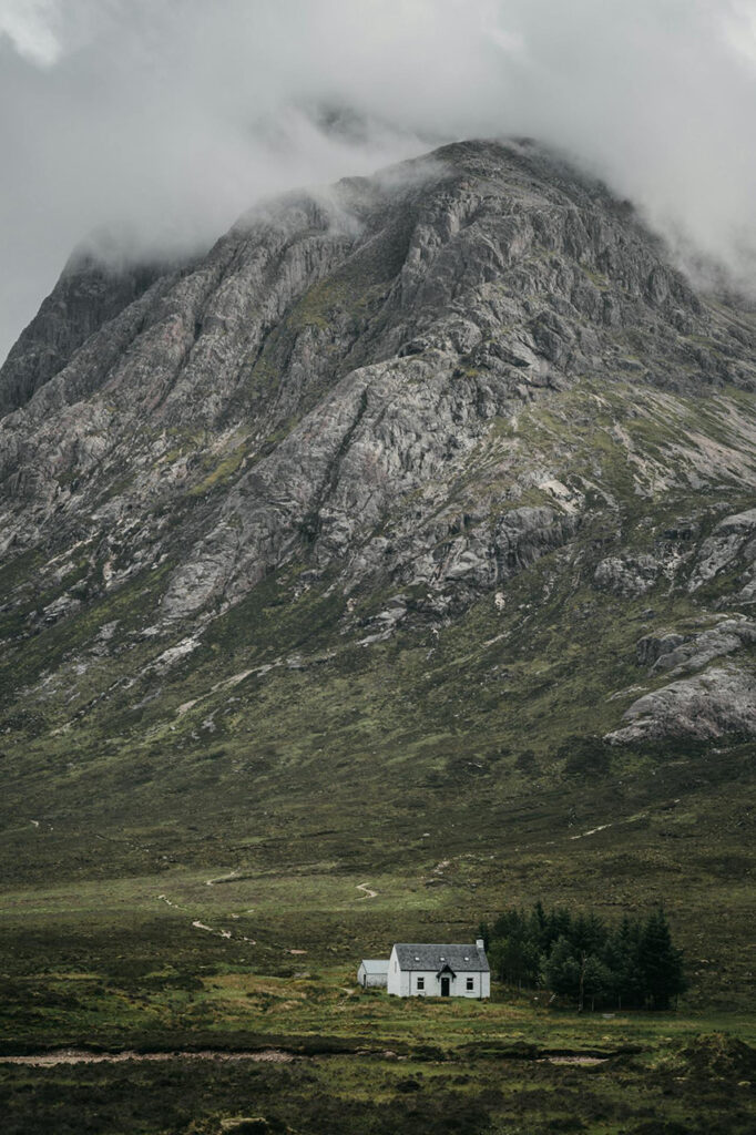 A small white house surrounded by greenery, set against the dramatic backdrop of Buachaille Etive Mor, a rugged mountain shrouded in mist, located in Glencoe, Scotland.