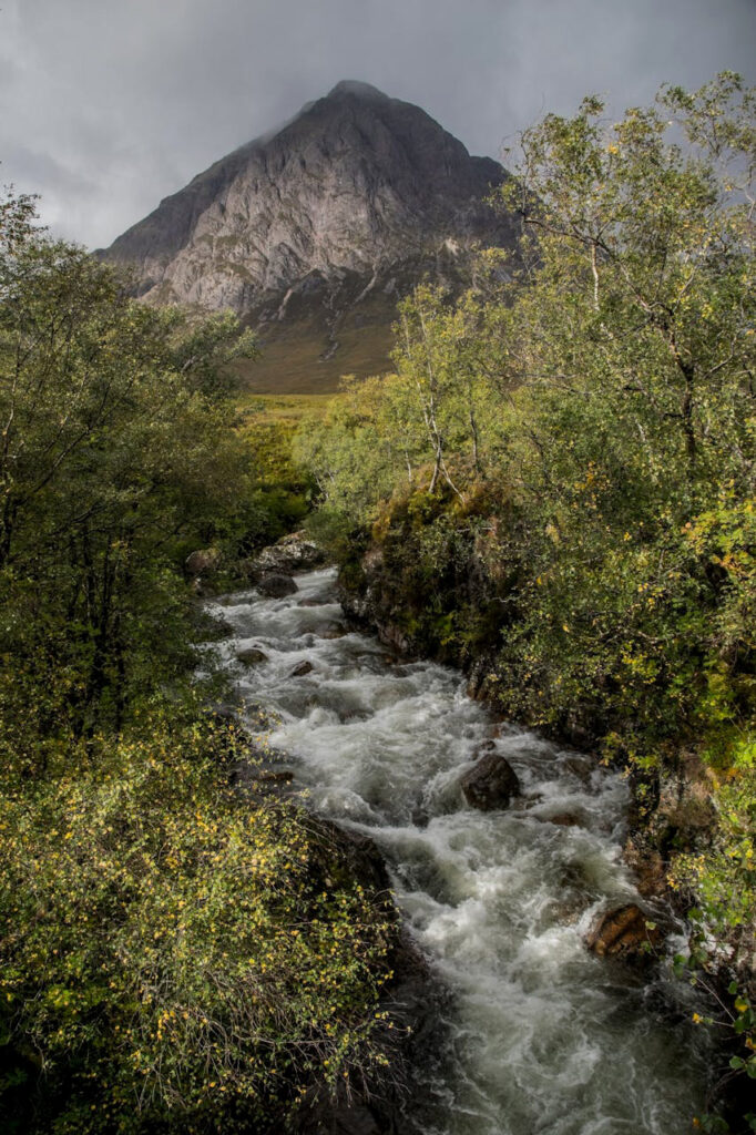 A scenic view of a rushing river surrounded by lush green trees, with the imposing Buachaille Etive Mor mountain rising in the background under a cloudy sky in Glencoe, Scotland.