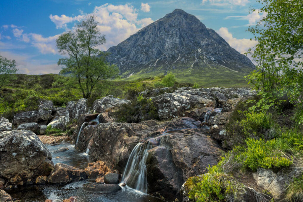 A tranquil small waterfall cascading over rocks, surrounded by lush greenery, with the iconic Buachaille Etive Mor mountain standing tall in the background under a partly cloudy blue sky in Glencoe, Scotland.