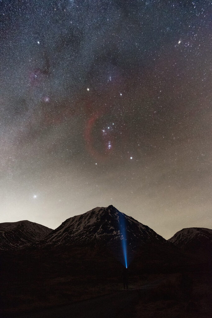 A starry night sky over Glencoe, with constellations vividly visible. A person shines a bright beam of light toward the snowy peak of a mountain, creating a dramatic silhouette against the cosmic backdrop.