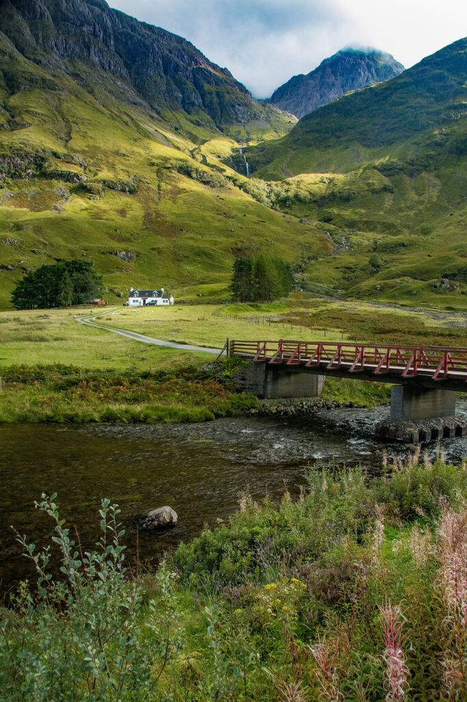 A picturesque scene in Glencoe, featuring a small red bridge crossing a clear river, with a white cottage nestled among vibrant green hills in the background, under a partly cloudy sky.