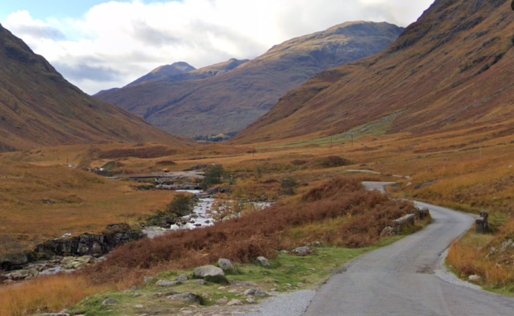 A winding single-track road snakes through the dramatic, rugged valley of Glencoe, surrounded by towering mountains and vast golden-brown moorland, with a stream meandering through the landscape. This iconic location, known as "Skyfall Glencoe," served as a backdrop for a pivotal scene in the James Bond movie Skyfall.