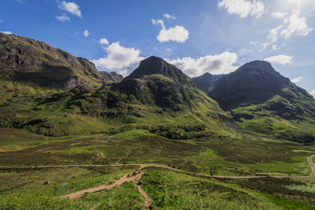 A breathtaking view of the Three Sisters of Glencoe, a trio of dramatic, steep-sided ridges surrounded by lush green landscapes under a vibrant blue sky dotted with fluffy white clouds.